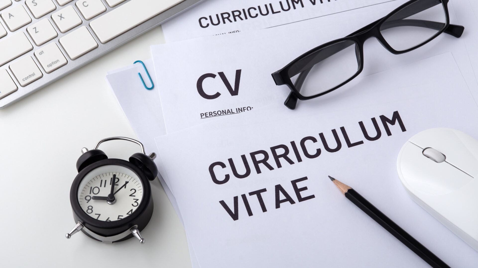 L'immagine mostra una scrivania ordinata su cui sono posizionati diversi fogli con la scritta "CURRICULUM VITAE" e "PERSONAL INFO". Accanto ai documenti, ci sono un paio di occhiali neri, una matita affilata, un mouse bianco e un orologio a sveglia che indica l'ora. Una tastiera di computer è visibile sul lato sinistro dell'immagine. Questa configurazione suggerisce l'importanza della preparazione quando si invia un curriculum vitae, e la "lettera di presentazione" è un elemento chiave in questo processo, fungendo da complemento al CV per fornire ulteriori dettagli sulla candidatura di una persona